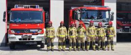 Zdjęcie przedstawia siedmiu strażaków ubranych w piaskowe munduru specjalne i czerwone hełmy stojących w rzędzie przed dwoma czerwonymi samochodami strażackimi.