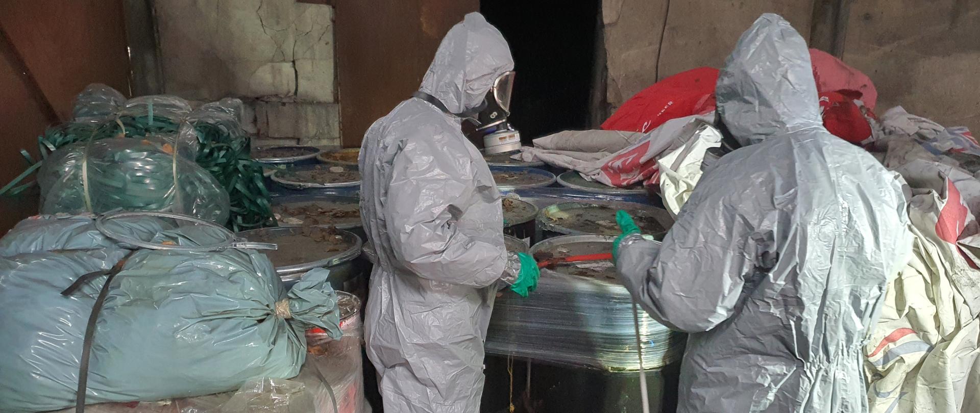 strażacy w ubraniach chemicznych oraz w sprzęcie ochrony dróg oddechowych stoja przes beczkami z subdtancją nieznanego pochodzenia