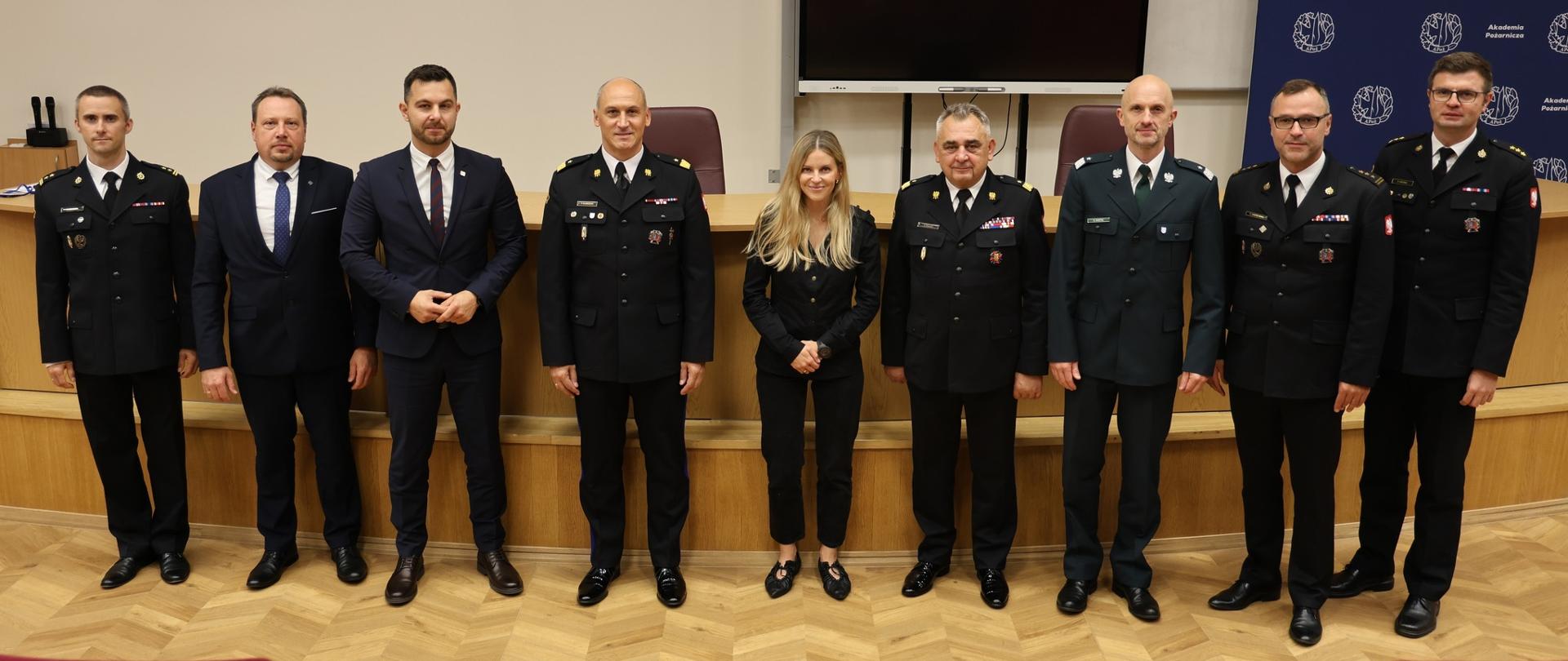 Grupa dziewięciu osób – przedstawicieli służb mundurowych i instytucji państwowych – pozuje wspólnie do zdjęcia na tle sali wykładowej. W centrum stoi kobieta w czarnym stroju, po bokach funkcjonariusze w mundurach PSP i innych formacji.