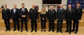 Grupa dziewięciu osób – przedstawicieli służb mundurowych i instytucji państwowych – pozuje wspólnie do zdjęcia na tle sali wykładowej. W centrum stoi kobieta w czarnym stroju, po bokach funkcjonariusze w mundurach PSP i innych formacji.