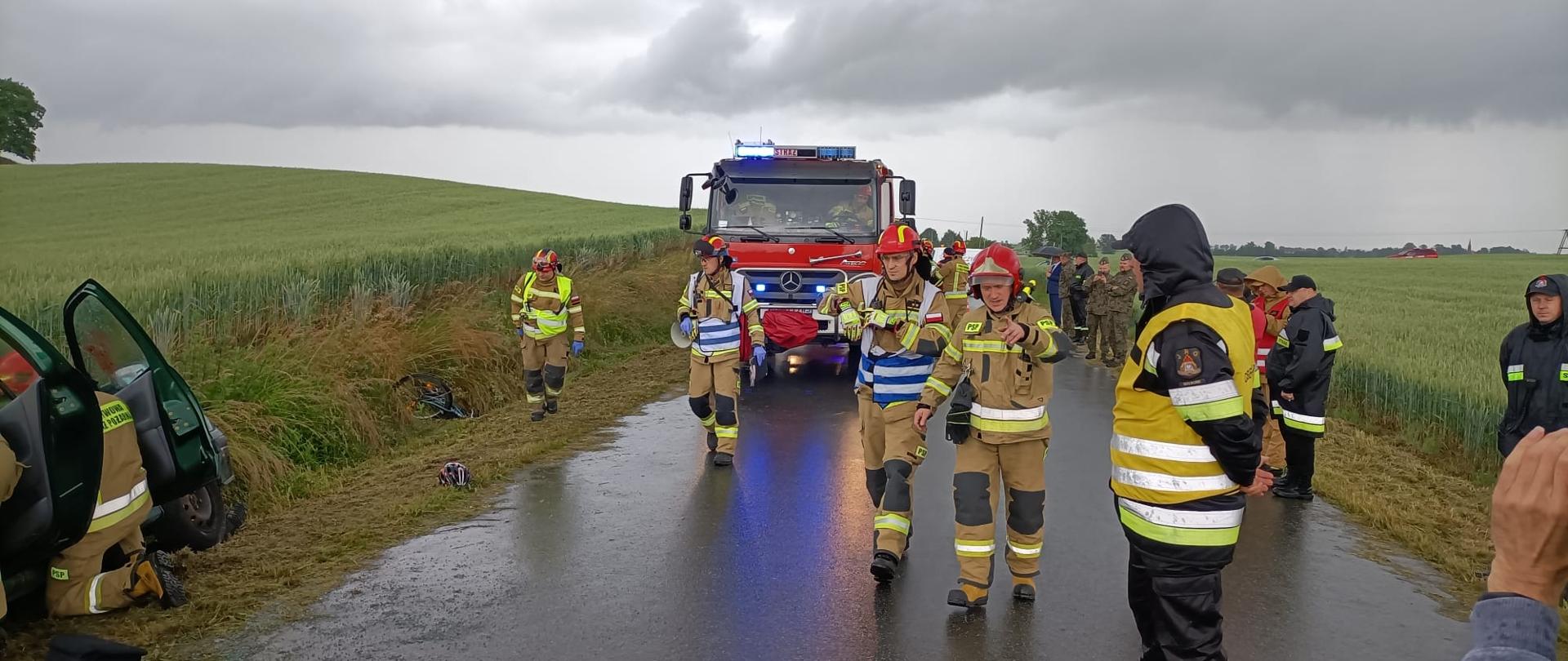 na drodze widać ustawiony pojazd pożarniczy przed którym idą do poszkodowanych strażacy.