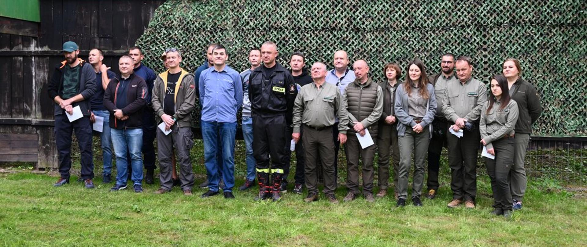 Kolorowa fotografia wykonana w pochmurny dzień na zewnątrz. Przestawia uczestników zawodów strzeleckich stojących podczas rozpoczęcia zawodów w dwuszeregu w strojach cywilnych i jeden strażak w ubraniu koszarowym.
