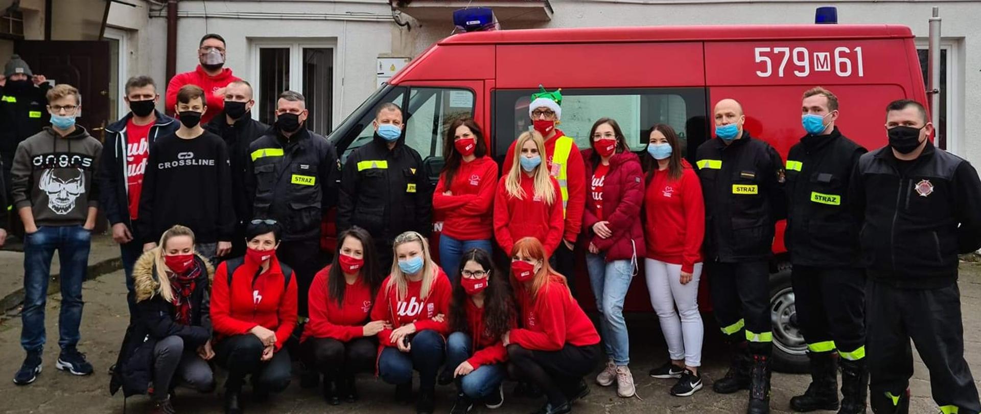 W środku grupa ludzi, część ubrana w czerwone struje, cz esc w ubrania strażackie, z tyłu samochód pożarniczy, w tle budynek Urzędu Gminy w Płońsku, murowany, jednokondygnacyjny.