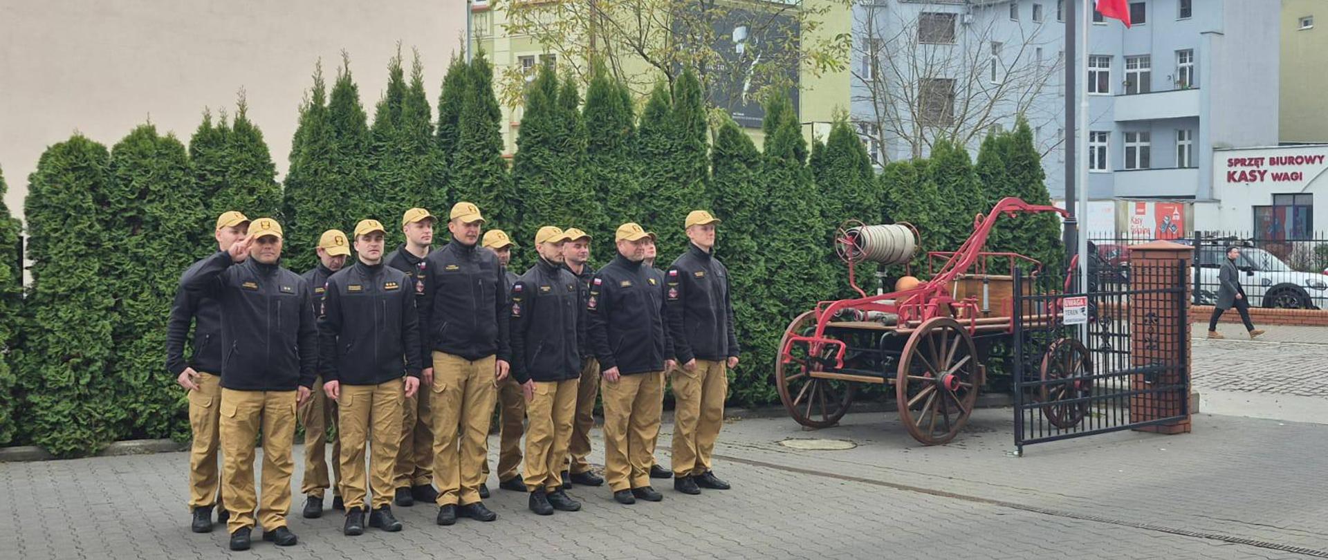 Zbiórka przy JRG1 Bydgoszcz na placu z szarej kostki, strażacy w czarnych polarach, żółtych spodniach i żółtych czapkach, w tle po prawej maszt z Flagą Państwową. 