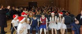 Na krzesłach w głębi sali zasiadają przedszkolaki. Mają ręce uniesione do góry. W pierwszym rzędzie siedzą uczniowie szkoły muzycznej w świątecznych strojach - czapkach mikołaja, białych spódniczkach i białych koszulkach.