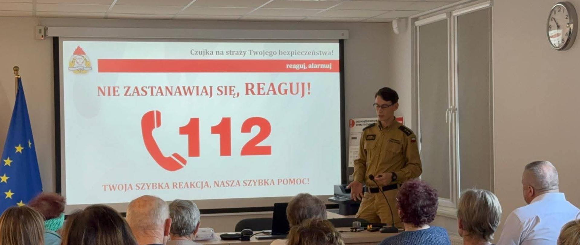 kpt. Maciej Wesserling przedstawia słuchaczom zasady powiadamiania służb ratunkowych. Na tablicy widnieje numer 112. 