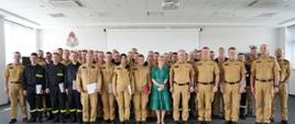 Zdjęcie grupowe uczestników szkolenia oraz członków kadry szkoleniowej stojących w dużym jasnym pomieszczeniu z szarą podłogą. Na ścianie za grupą osób widoczne jest logo Państwowej Straży Pożarnej oraz duży biały ekran.