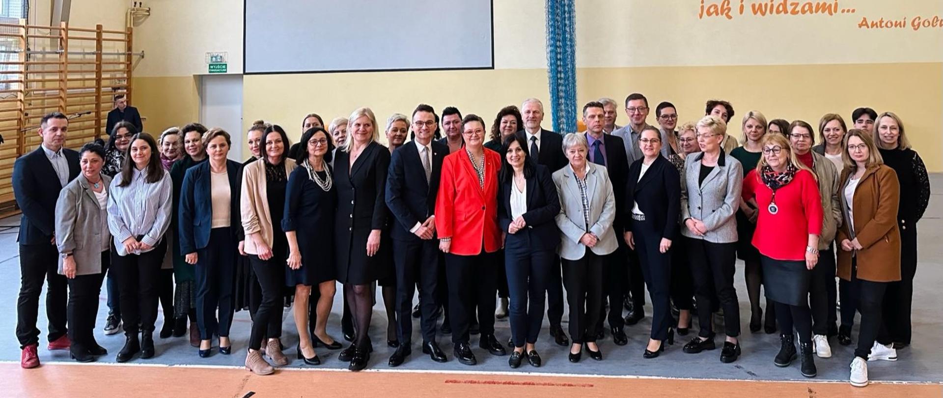 Zdjęcie zbiorowe, na sali pod ścianą stoi duża grupa ludzi. pomiędzy nimi wiceminister Katarzyna Lubnauer. W tle widać drabinki gimnastyczne i napis na ścianie: Życie ludzkie to olbrzymie zawody sportowe, których jesteśmy zarówno uczestnikami jak i widzami... Antoni Golubiew 