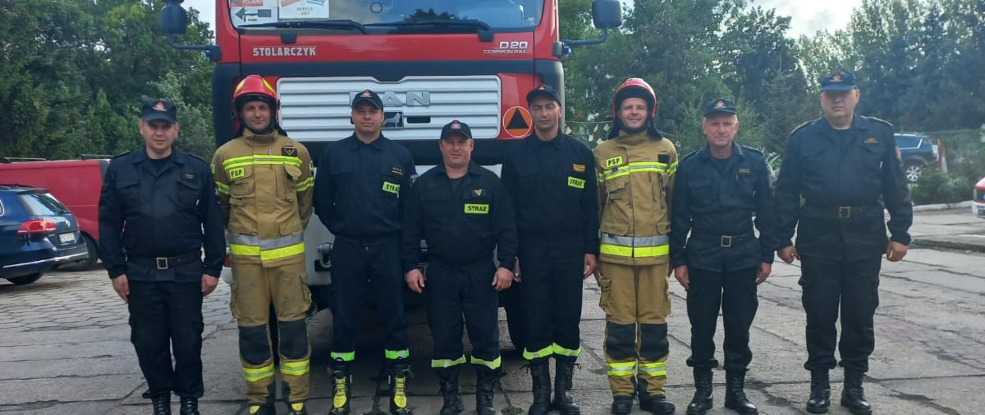 Na zdjęciu widać strażaków ubrane w czarne oraz piaskowe mundury. Strażacy stoją przed samochodem gaśniczym Man w kolorze czerwonym z sygnalizacją ostrzegawczą. Strażacy stoją na szarych płytach betonowych. Niebo lekko zachmurzone, w kolorze błękitnym.