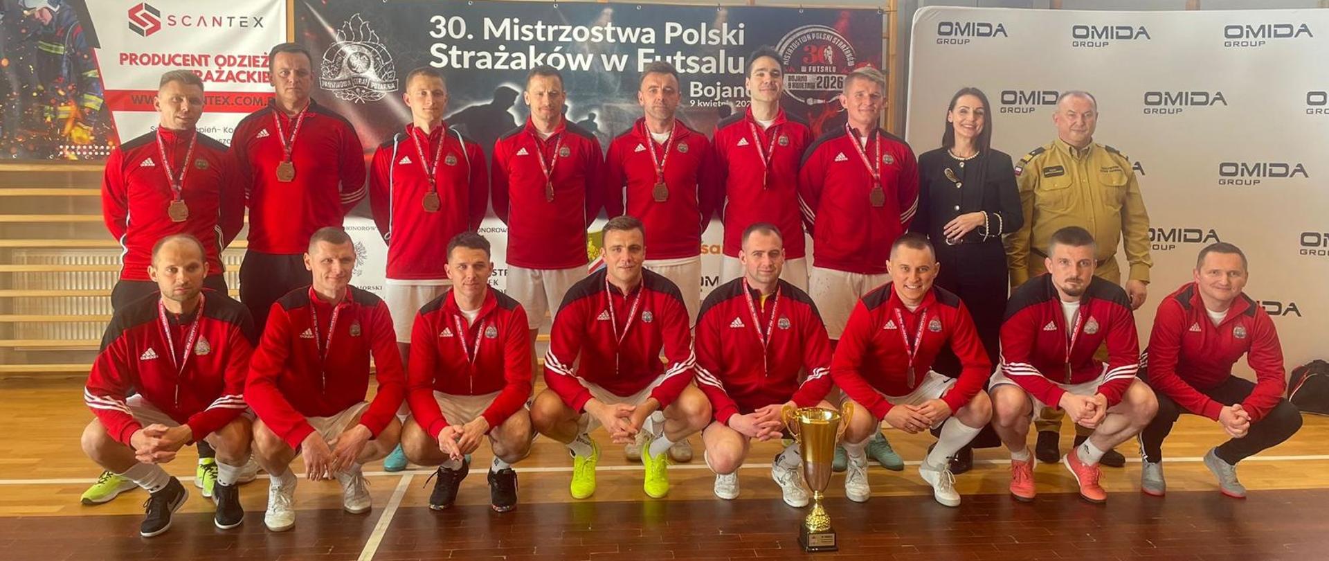 Zdjęcie przedstawia drużynę, która wygrała mecz w futsalu. Zdjęcie zrobione na hali sportowe. Mężczyźni ustawieni w dwóch rzędach. Drugi rząd w pozycji stojącej pierwszy w pozycji "kucającej" Panowie na sobie mają strój sportowy: spodenki i bluza.