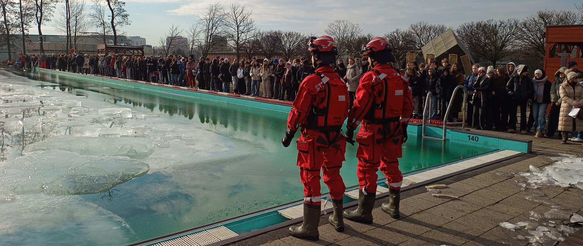 Zdjęcie przedstawia zimowe wydarzenie plenerowe odbywające się przy dużym, odkrytym basenie wypełnionym zielonkawą wodą i krą lodową. Na pierwszym planie, tyłem do obiektywu, stoją dwaj strażacy w pełnym, czerwonym rynsztunku – mają na sobie specjalistyczne kombinezony wodne, kaski oraz kamizelki asekuracyjne. Ich obecność sugeruje, że pilnują bezpieczeństwa podczas pokazów ratownictwa.
Wzdłuż krawędzi basenu zgromadził się liczny tłum gapiów, ubranych w ciepłe, zimowe kurtki i czapki. Ludzie z zaciekawieniem obserwują taflę wody, a w tle widać bezlistne drzewa oraz zabudowania miejskie pod jasnym, lekko zachmurzonym niebem. Cała scena ma charakter edukacyjny lub sportowy, typowy dla miejskich akcji promujących bezpieczeństwo nad wodą zimą.
