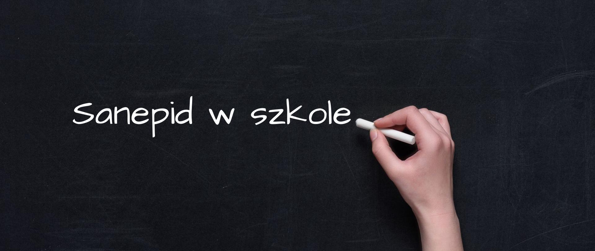 Na zdjęciu jest tablica szkolna, na której jest napisane sanepid w szkole. Widoczna jest ręka, w której znajduje się kreda.