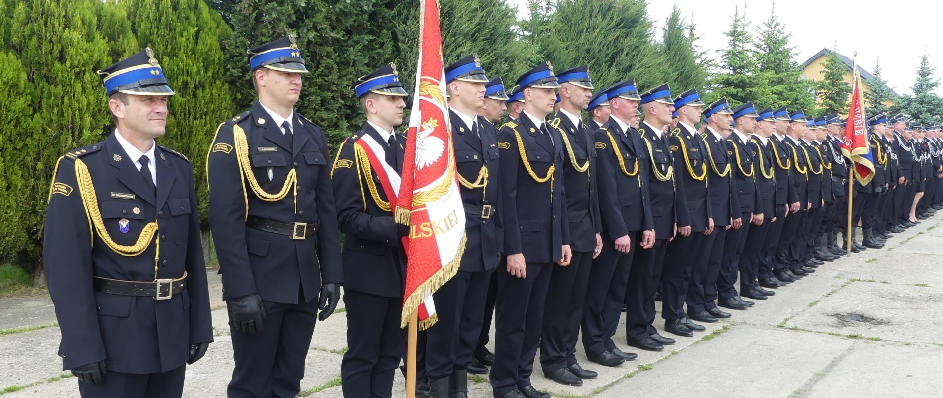 Zdjęcie przedstawia strażaków w mundurach galowych. Po lewo stoi Dca JRG w Brodnicy. Obok niego – po prawo stoi poczet sztandarowy ze sztandarem Komendy. Dalej po prawo w dwuszeregu strażacy KP PSP w Brodnicy. W oddali druhowie Jednostek OSP.