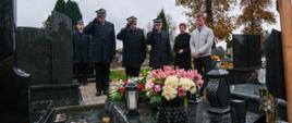 Strażacy Państwowej Straży Pożarnej w galowych mundurach oddają honory przy grobie zmarłego funkcjonariusza. Przed nagrobkiem stoją znicze i bukiety kwiatów. W tle widoczne są drzewa z jesiennymi liśćmi i inne groby na cmentarzu.