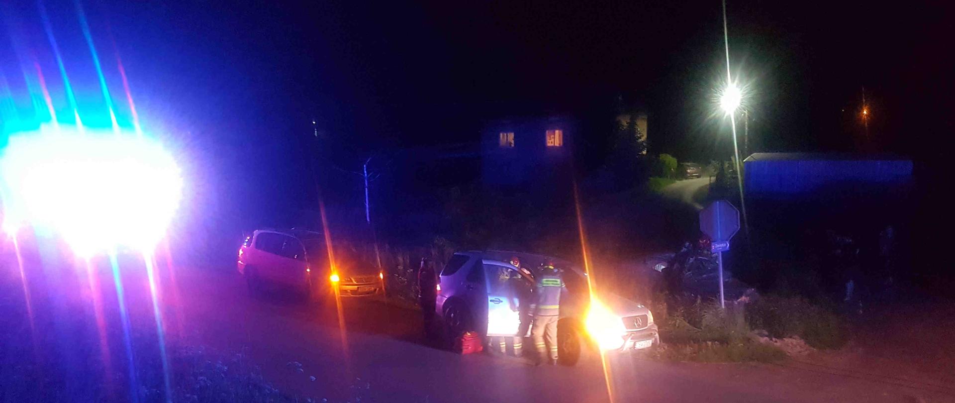 Na pierwszym planie ratownicy udzielają kpp poszkodowanemu kierowcy, który został ewakuowany do samochodu osobowego jednego ze świadków zdarzenia. Z lewej strony widać samochód ratowniczo-gaśniczy a na ostatnim planie samochód, który uległ wypadkowi