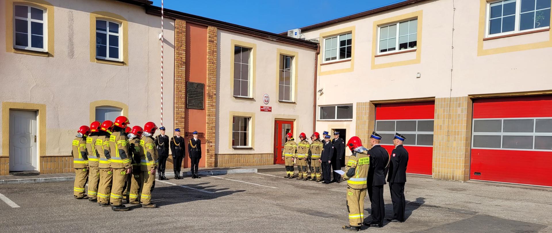 Zdjęcie przedstawia zbiórkę strażaków stojących w czworokącie na placu między budynkami. Większość z nich ubrana jest w piaskowe mundury bojowe i czerwone hełmy pozostali mają mundury galowe i rogatywki. W tle czerwone bramy okna i drzwi budynku.