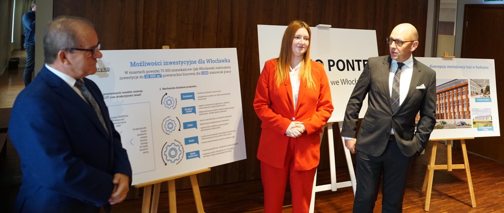 wiceminister Anna Gembicka i inni uczestnicy spotkania stoją i pokazują na plansze z prezentacją programu Fabryka