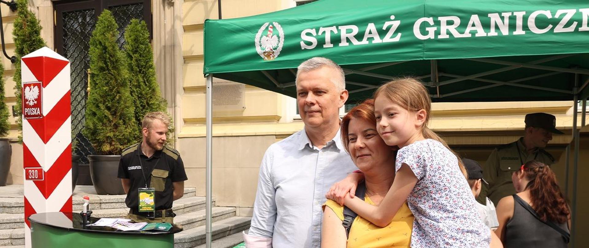 Minister Tomasz Siemoniak, obok mama z dziewczynką na rękach. W tle namiot straży granicznej 