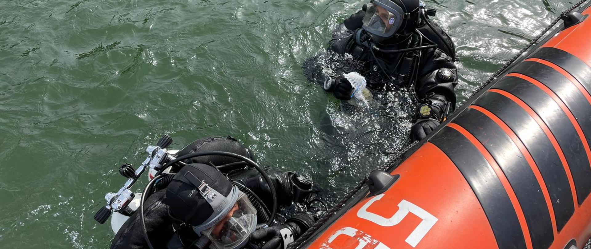 Dwóch nurków jest w wodzie przy pontonie. Przygotowani są do zejścia pod wodę. 