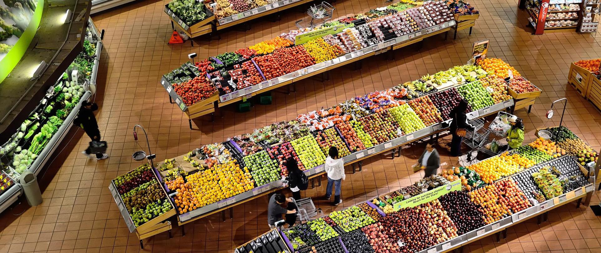 Na zdjęciu znajduje się (widok z góry) dział warzyw i owoców w supermarkecie - stoją rzędy wyłożonych na stojakach świeżych owoców i warzyw.