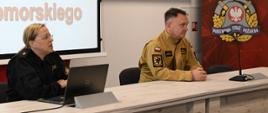 Za stołem siedzą funkcjonariusz w czarnym mundurze przed laptopem, obok strażak w mundurze w kolorze piaskowym. Obok baner Państwowej Straży Pożarnej.