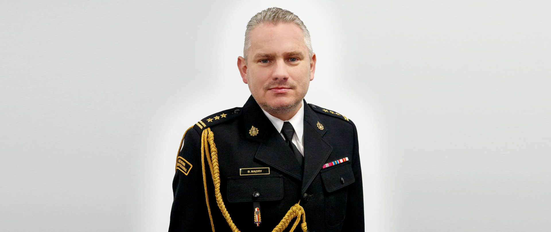 Zdjęcie przedstawia st. bryg. Bartłomieja Mądrego Zastępcę Lubuskiego Komendanta Wojewódzkiego PSP w Gorzowie Wlkp. w mundurze na szarym tle. 