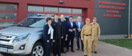 Przekazanie samochodu na placu komendy, Komendant Powiatowy Państwowej Straży Pożarnej w Brzezinach starszy brygadier Mariusz Kaczmarski prezes jednostki Ochotniczej Straży Pożarnej w Woli Cyrusowej druh Jarosławowi Piórkowskiemu, Bartłomiej Lipski – Starosta Brzeziński, Arkadiusz Garnys – Wicestarosta Brzeziński, Danuta Supera – Wójt Gminy Dmosin, mł. bryg. Jakub Brzózka – Zastępca Komendanta Powiatowego Państwowej Straży Pożarnej w Brzezinach, druh Piotr Wiśniewski – wiceprezes jednostki OSP w Woli Cyrusowej, pozują do zdjęcia podczas uroczystego przekazaniu samochodu. Stoją obok samochodu za samochodem i pozującymi widać budynek komendy.