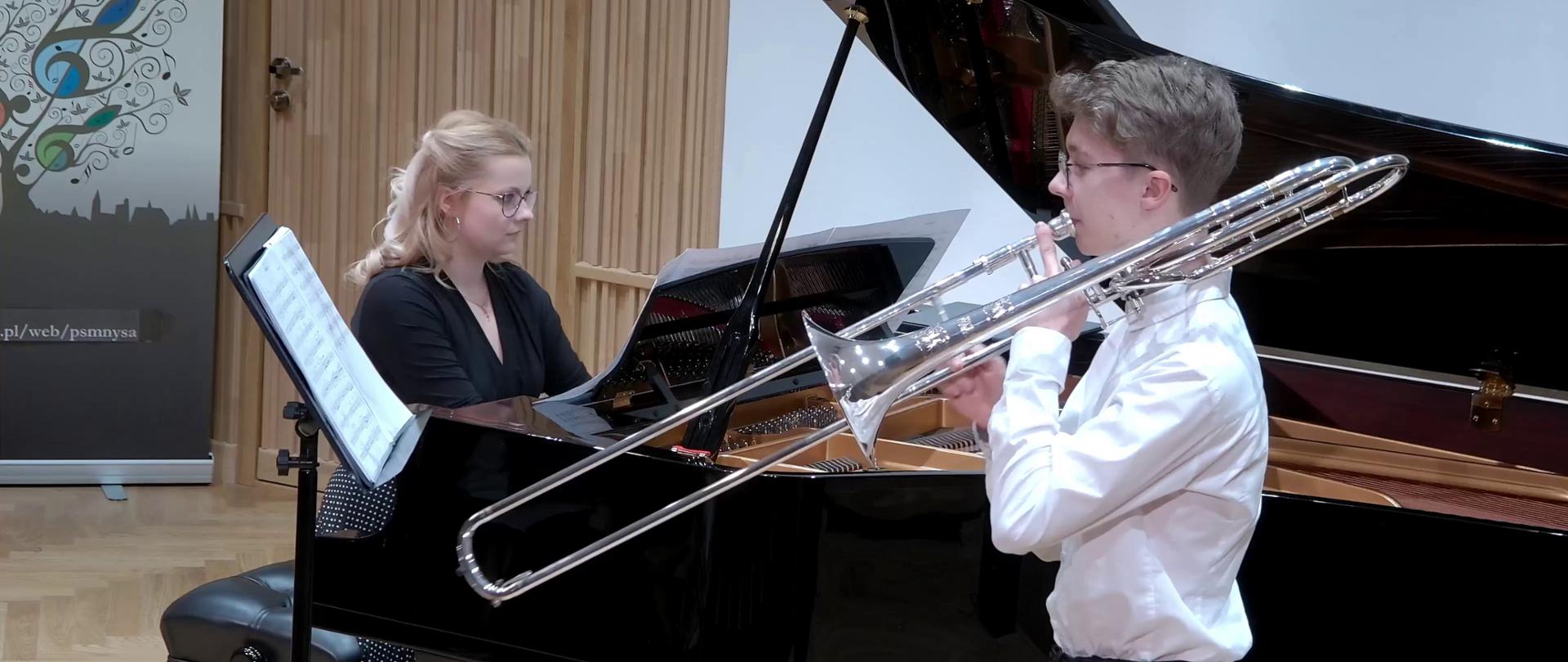 Zdjęcie. Na estradzie występuje młody puzonista z towarzyszeniem pianistki, która gra na dużym czarnym fortepianie. Przed puzonistą stoi pulpit z nutami. W głębi w tle z lewej strony widać fragment baneru PSM Nysa.
