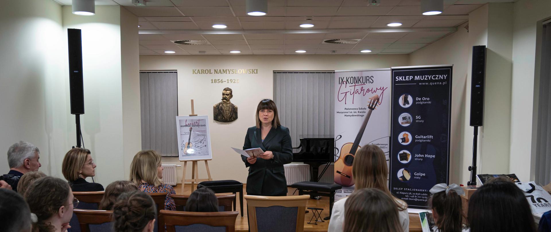 Sala koncertowa Państwowej Szkoły Muzycznej I st. im. K. Namysłowskiego w Szczebrzeszynie. Na zakończenie IX Konkursu Gitarowego, przed koncertem Kupiński Guitar Duo, Iwona Grabowska przedstawia sylwetki wykonawców. Stoi po środku sali, przed sceną, ubrana w zieloną marynarkę, trzyma w dłoni program. Przed nią siedzą ludzie na krzesłach - widownia ujęta plecami.
