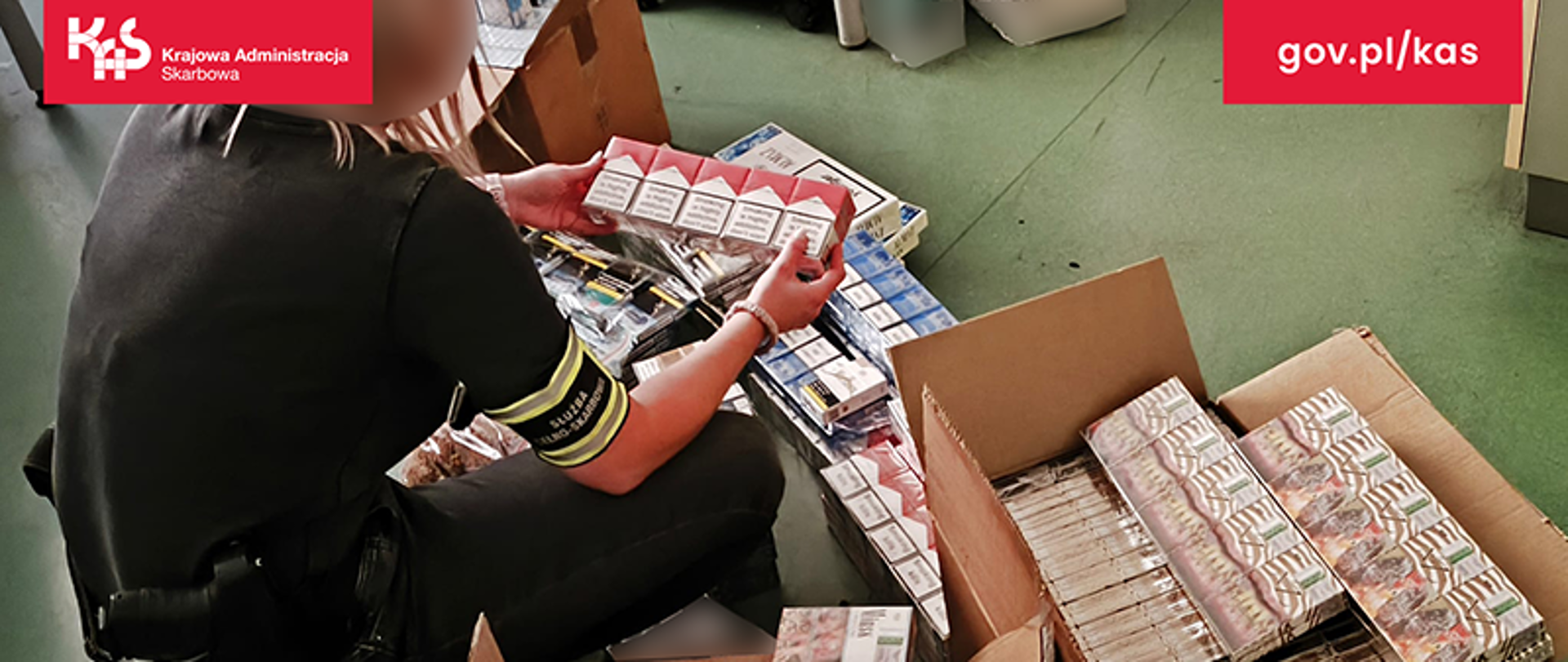 Na podłodze leży duża ilość papierosów różnych marek. Przy wyrobach tytoniowych kuca funkcjonariusz Urzędu Celno-Skarbowego.