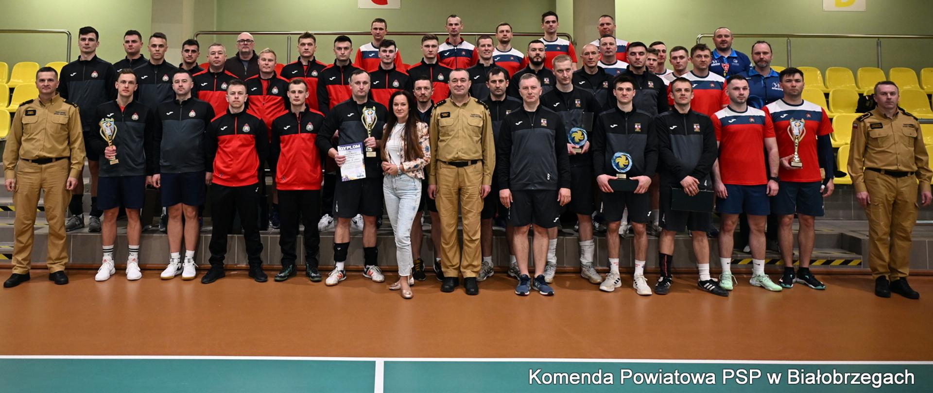 Duże zdjęcie grupowe wszystkich uczestników turnieju, sędziów i organizatorów na tle trybun. W pierwszym rzędzie stoją kapitanowie z pucharami i dyplomami oraz funkcjonariusze w mundurach służbowych. Nad nimi, na barierkach trybun, wisi baner charytatywny LEON SŁOMKA Uratuj Życie