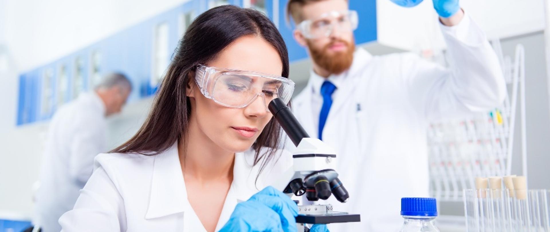 Team work. Young lab worker in safety glasses is analysing the sample in the microscope. She is in a labcoat, in the middle of experiment