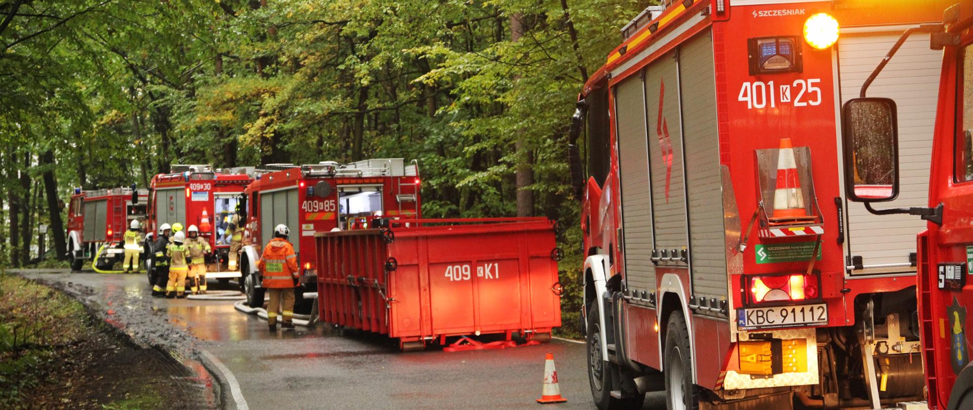 Wozy strażackie zaparkowane po prawej stronie jezdni na asfaltowej drodze w lesie pośrodku kolumny stoi kontener w umaszczeniu czerwonym w dalszej części kolumny obok wozów stoją strażacy w pomniejszych grupkach
