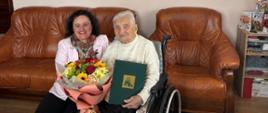 Uśmiechnięta starsza Pani na wózku inwalidzkim trzyma w rękach zieloną teczkę ze złotym logo KRUS z listem gratulacyjnym Prezesa KRUS, po jej prawej stronie przykucnęła młodsza kobieta trzymająca w rękach bukiet kwiatów.