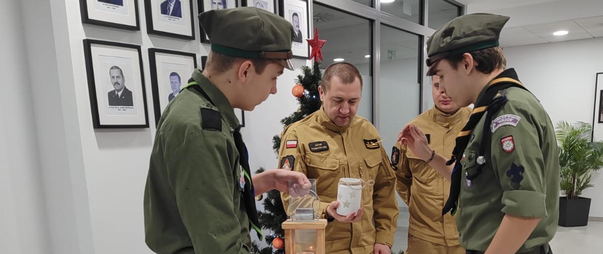 Zdjęcie przedstawia dwóch strażaków i dwóch harcerzy przekazujących światło pokoju