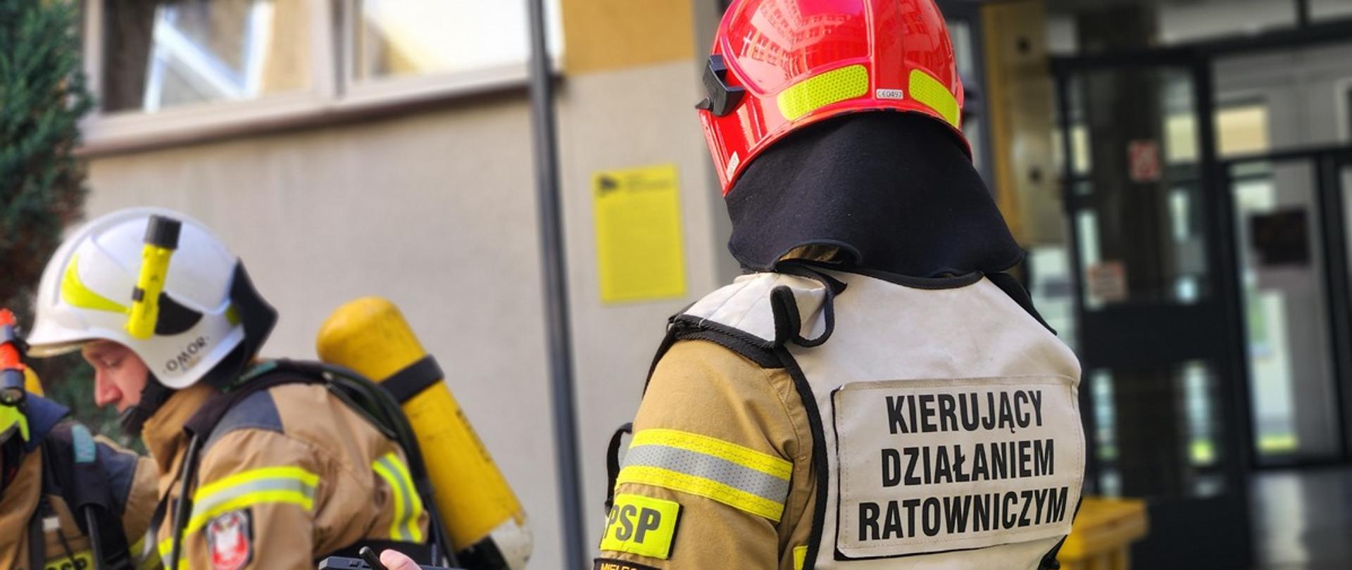 Portret strażaka od tyłu, ubranego w kamizelkę z napisem „KIERUJĄCY DZIAŁANIEM RATOWNICZYM”. W ręku trzyma radiostację. W tle widoczni inni strażacy przygotowujący sprzęt oraz wejście do budynku szkoły.