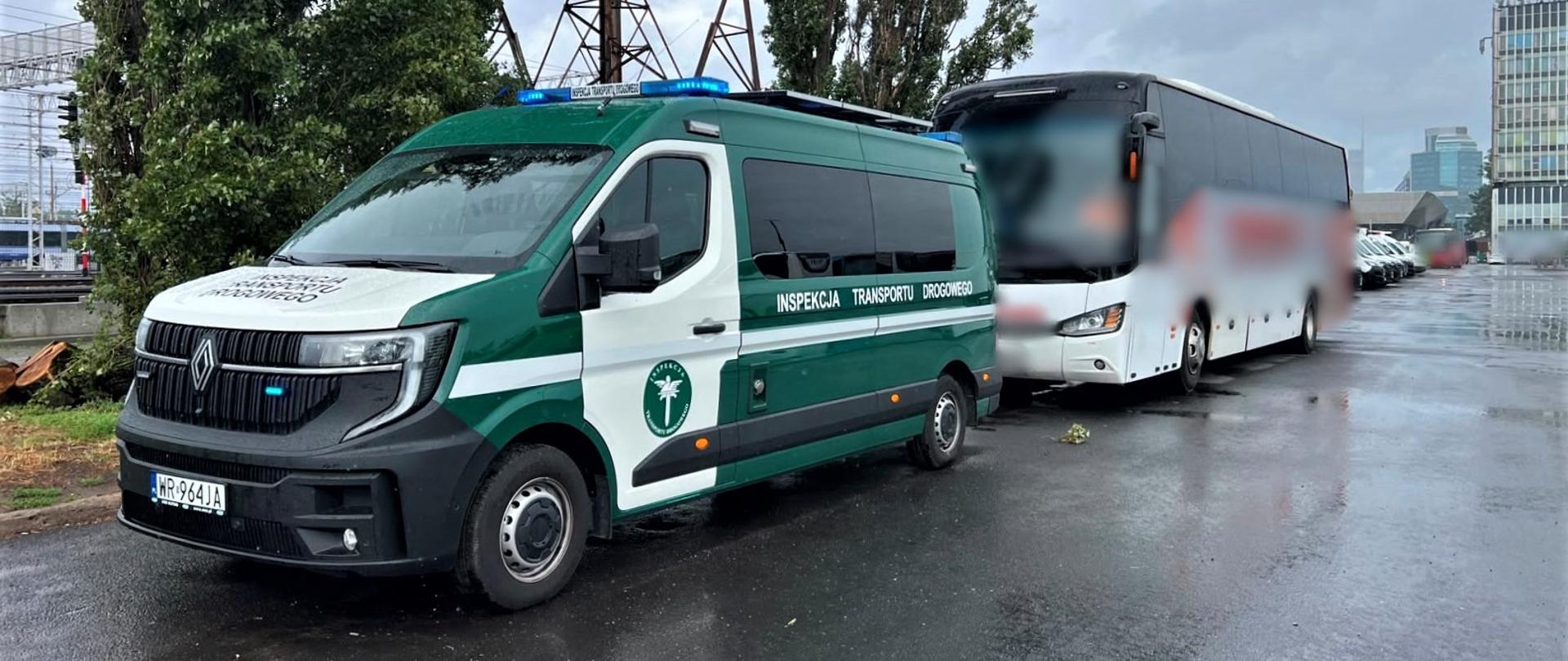 Na pierwszym planie (od lewej): przód i lewy bok oznakowanego furgonu mazowieckiej Inspekcji Transportu Drogowego. Radiowóz ma włączone niebieskie światła uprzywilejowania w ruchu drogowym. W tle: za furgonem inspekcyjnym stoi kontrolowany autokar. W oddali widać tory kolejowe i wysokie budynki biurowców w centrum Warszawy.