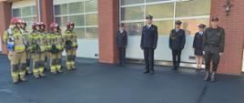 Przed garażami strażnicy stoją strażacy ubrani w mundurach specjalnych koloru beżowego oraz w mundurach wyjściowych.