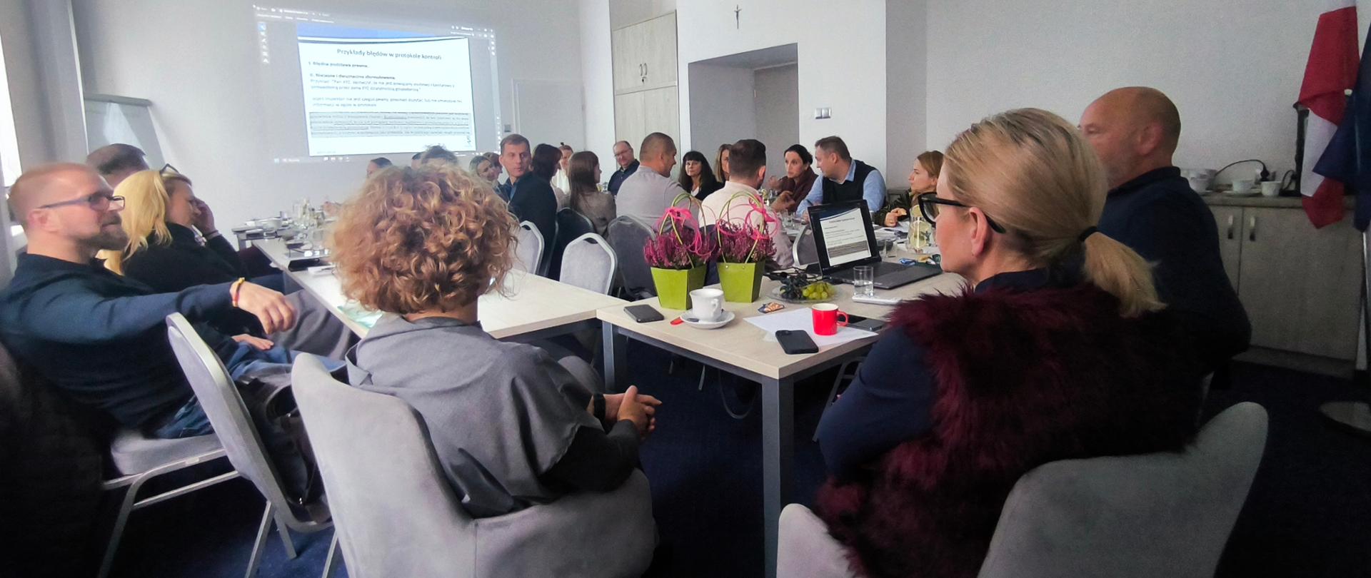 grupa osób siedzi przy stole, na ścianie wyświetlany jest slajd