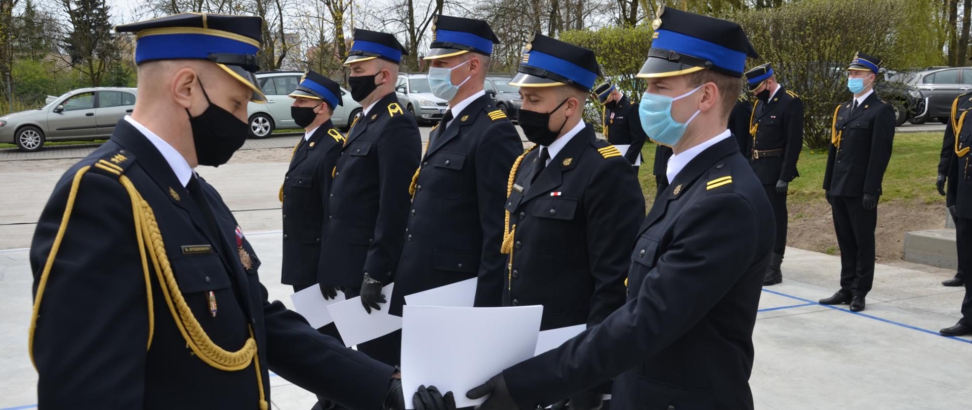 Na terenie Komendy Powiatowej Państwowej Straży Pożarnej odbywa się uroczystość obchodów Dnia Strażaka. Komendant Powiatowy w mundurze galowym wręcza awanse strażakom stojącym w szyku. Strażacy, którzy również ubrani są w mundury galowe odbierają wręczane awanse w wersji papierowej z rąk Komendanta Powiatowego.