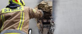 Na zdjęciu widoczny strażak na drabinie przy elewacji budynku. W elewacji budynku widoczna dziura po spalonej skrzynki elektrycznej i elementy wełny ocieplenia budynku.