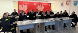 Na oświetlonej sali siedzą kobiety i mężczyźni ubrani w mundury straży pożarnej, w tle widoczne czerwone banery Państwowej Straży Pożarnej
