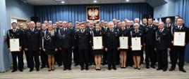 Zdjęcie wykonane w siedzibie komendy wojewódzkiej Państwowej Straży Pożarnej w Warszawie. Zdjęcie wykonane w formie zdjęcia pamiątkowego upamiętniającego uroczystość. Na zdjęciu widać ustawionych w rzędach kadrę kierowniczą komendy wojewódzkiej w towarzystwie przedstawicieli jednostek ochotniczych straży pożarnych które otrzymały akty włączenia. Zebrani ubrani są w mundury galowe w kolorze ciemnym granatowym. Ponadto przedstawiciele jednostek trzymają w rękach pamiątkowe akty.