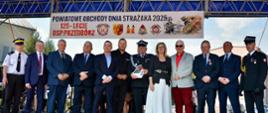 Zdjęcie grupowe osób w eleganckich strojach – mundurach strażackich oraz garniturach – stojących na scenie z niebieskim dachem. Za nimi transparent z napisem „Powiatowe Obchody Dnia Strażaka 2025 - 125-lecie OSP Przedbórz”.
