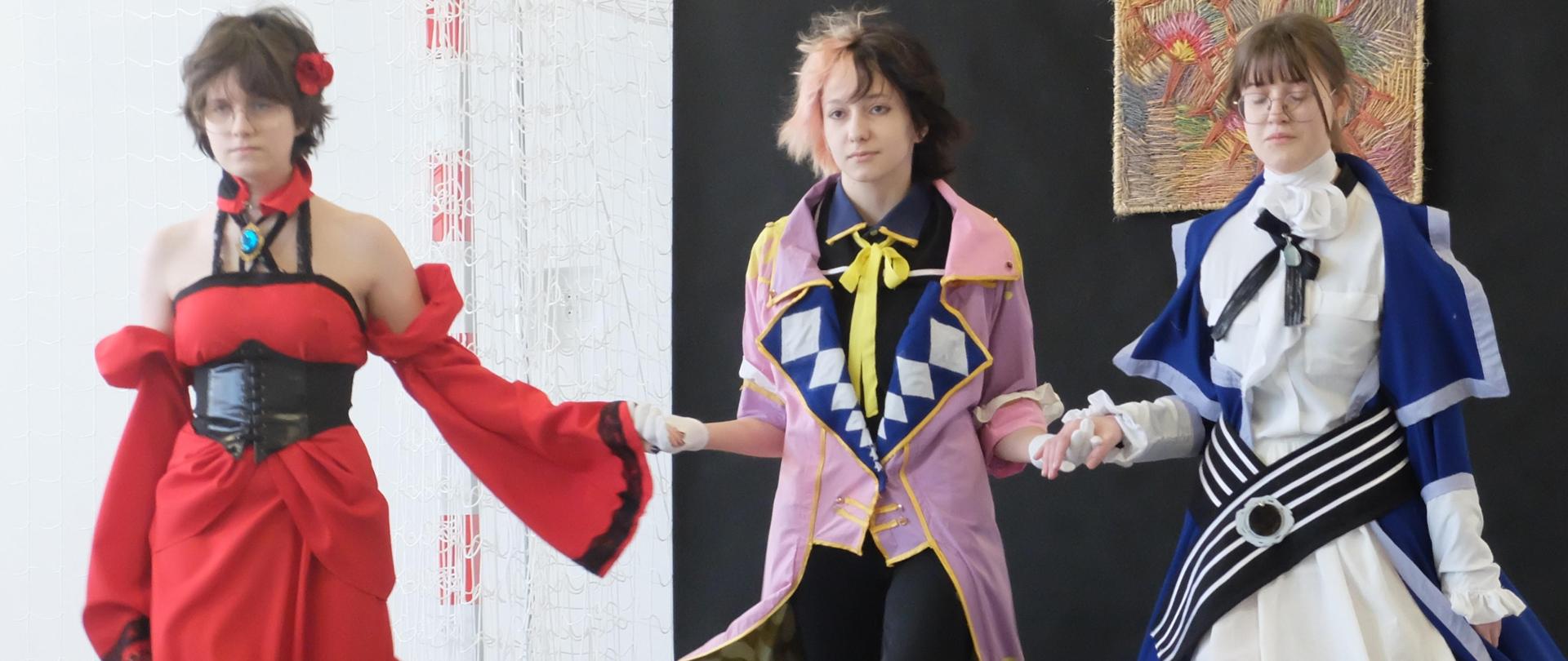 Trzy modelki w strojach inspirowanych grami i światem fantasy idą w pokazie na tle czarnej draperii. 