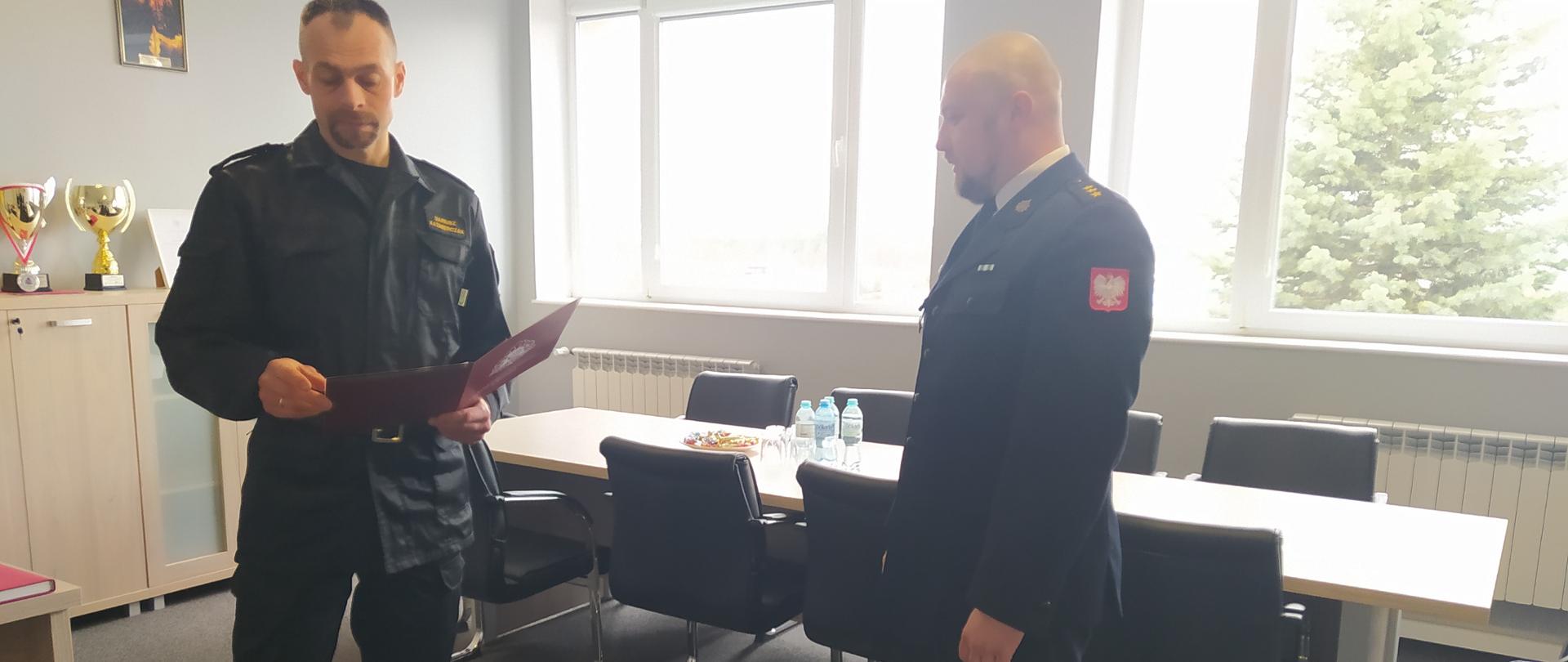 Po lewej stronie znajduje się Komendant KP PSP Bytów z czerwoną teczką w rękach. Po jego prawej stronie stoi na baczność strażak. Strażacy znajdują się w biurze. W tle widać stół konferencyjny oraz wyposażenie biurowe.