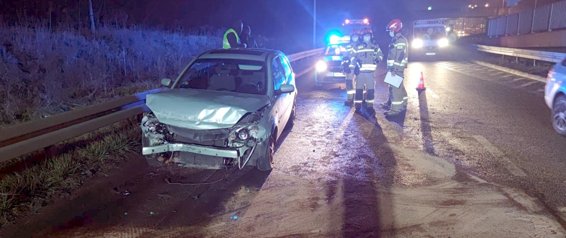 Zdjęcie przedstawia samochód osobowy marki Opel Astra ze zgniecionym przodem, stojący na poboczu drogi .Obok niego stoją strażacy i policjanci wykonujący czynności służbowe. W tle stoją samochody ratowniczo - gaśnicze oraz radiowóz policji z włączonymi światłami ostrzegawczymi. Z prawej strony zdjęcia widoczne są samochody przejeżdżające obok zdarzenia. 