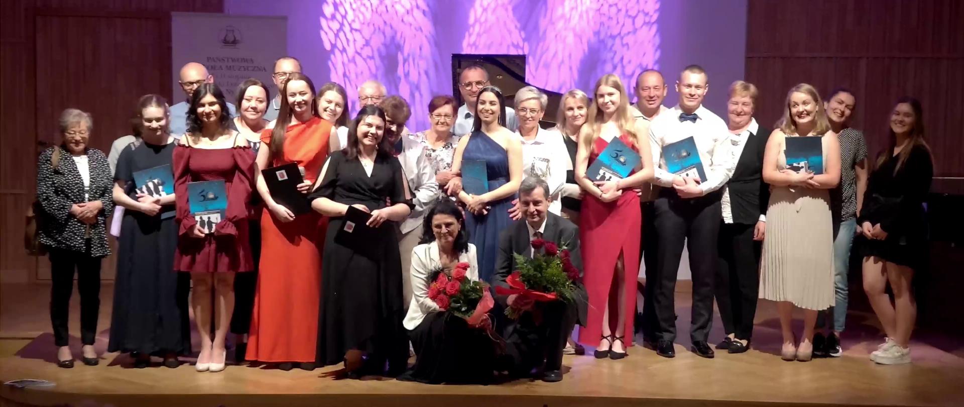 Zdjęcie. Na estradzie Sali Koncertowej stoją absolwenci szkoły wraz z nauczycielami i osobami bliskimi. W tle wyświetlona jest barwna grafika, obok widać fragment logo PSM Nysa. Absolwenci trzymają teczki z dyplomami, dyrekcja trzymając kwiaty przyklęka z przodu.