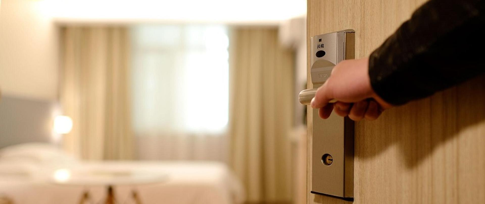 na pierwszym planie ręka na klamce drzwi, które zostały otworzone do pokoju hotelowego, na drugim planie stolik i łózko hotelowe, w tle okno