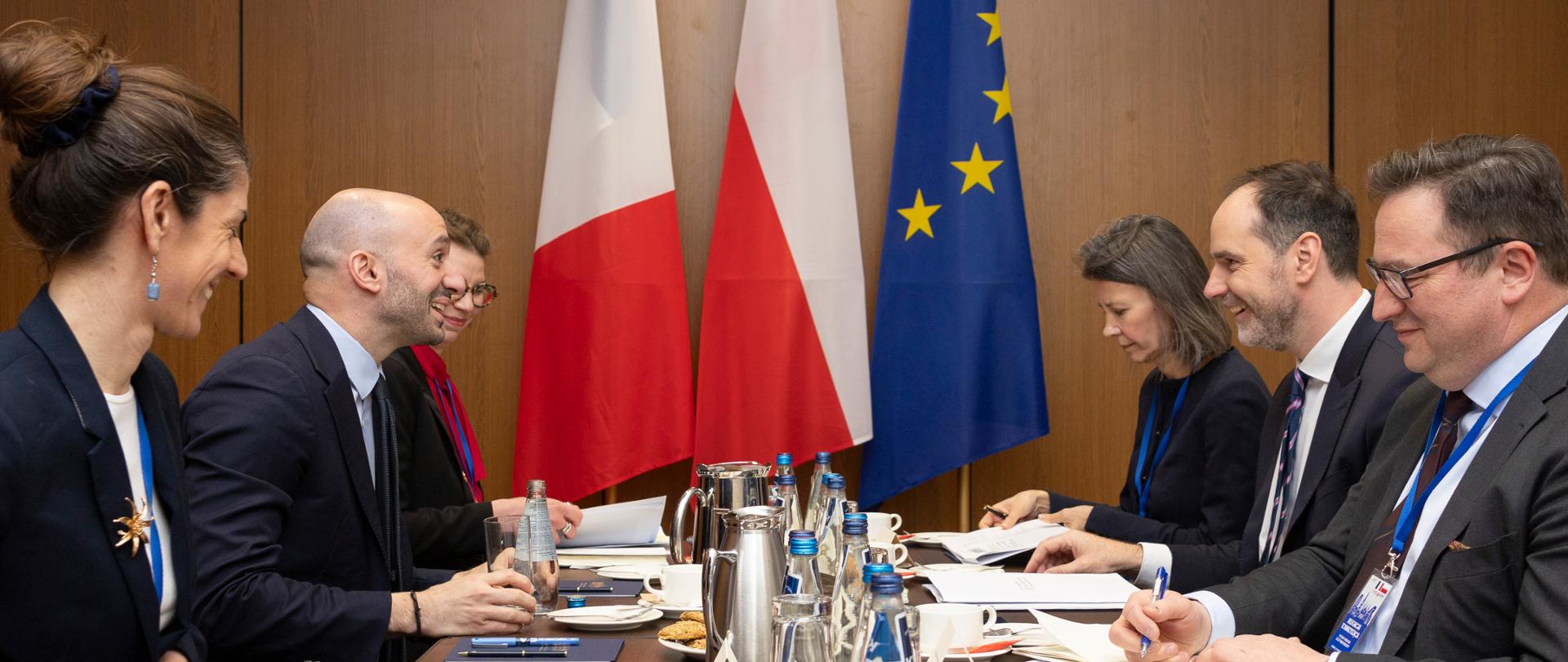 Delegations of France and Poland sit at a conference table during a meeting, talking in the presence of the flags of France, Poland, and the European Union. On the left: French Minister Delegate for Europe Benjamin Haddad and two women, on the right Secretary of State at the Polish Ministry of Foreign Affairs Ignacy Niemczycki and a man and a woman